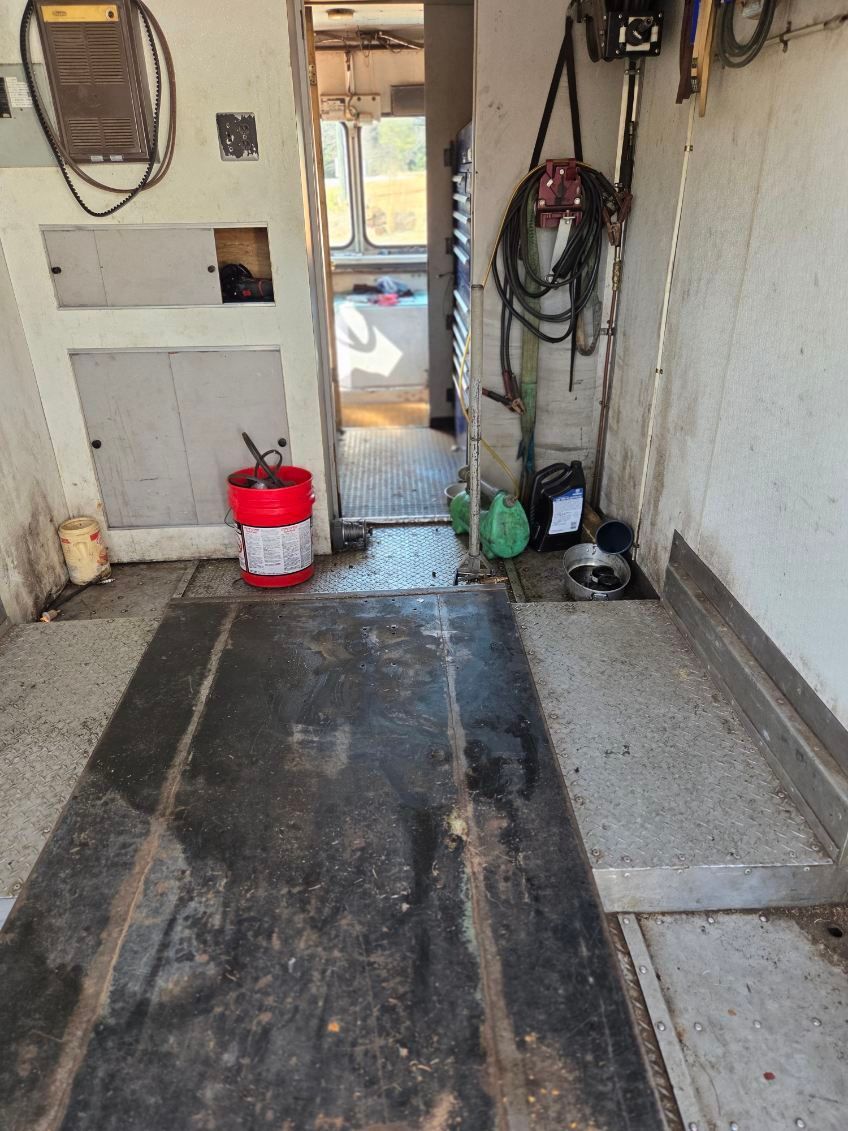 Narrow utility hallway with a worn black floor mat, red bucket, and hanging tools by a doorway.