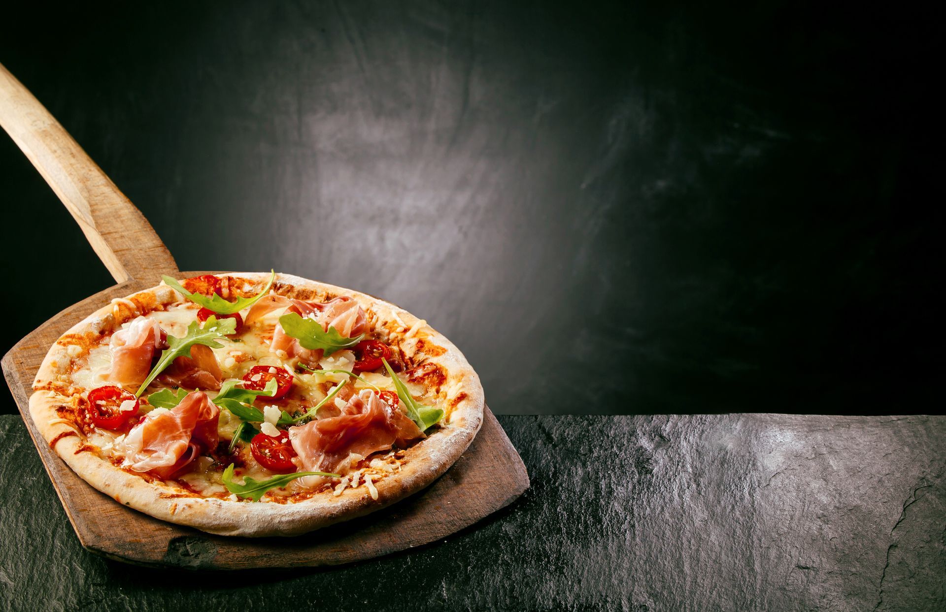 Prosciutto pizza topped with arugula, tomatoes, and cheese, served on a wooden pizza peel.