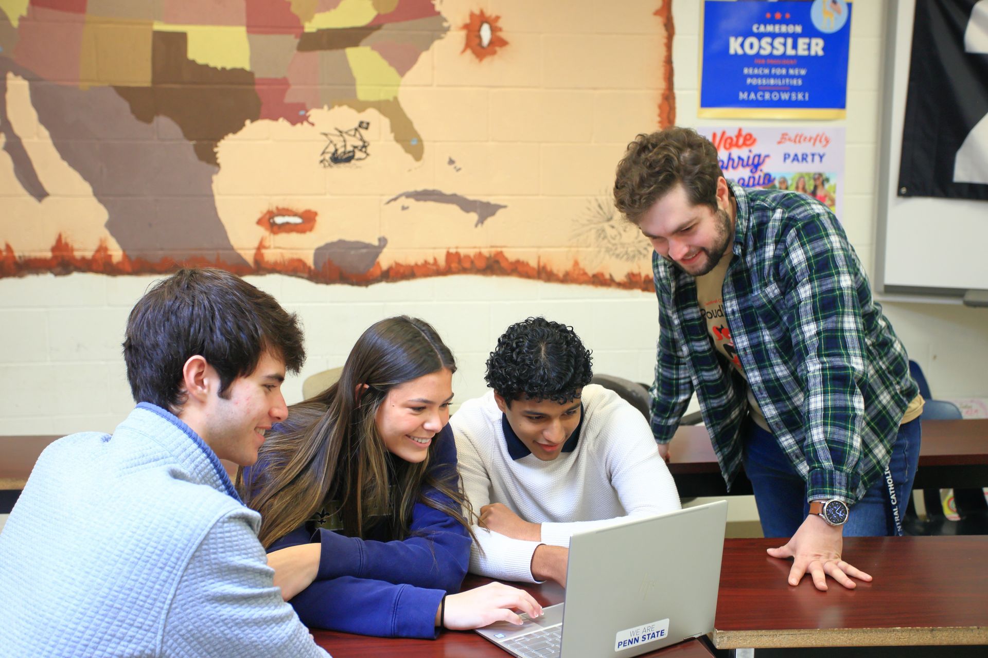 A group of people looking at a laptop with a poster on the wall that says kossler