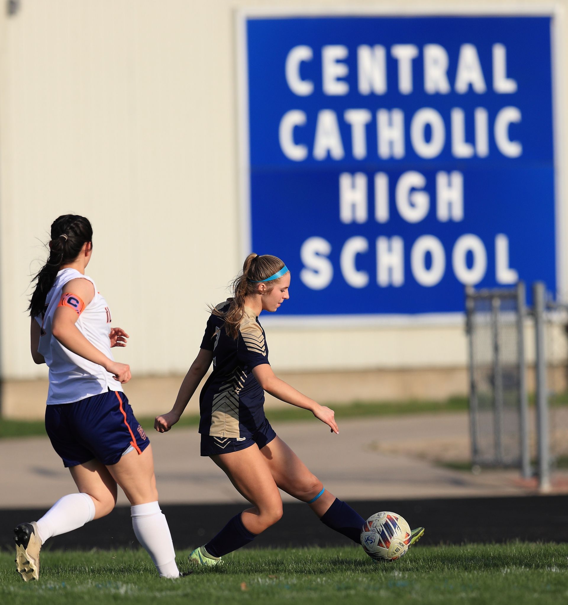 Two girls playing soccer in front of a sign that says central catholic high school