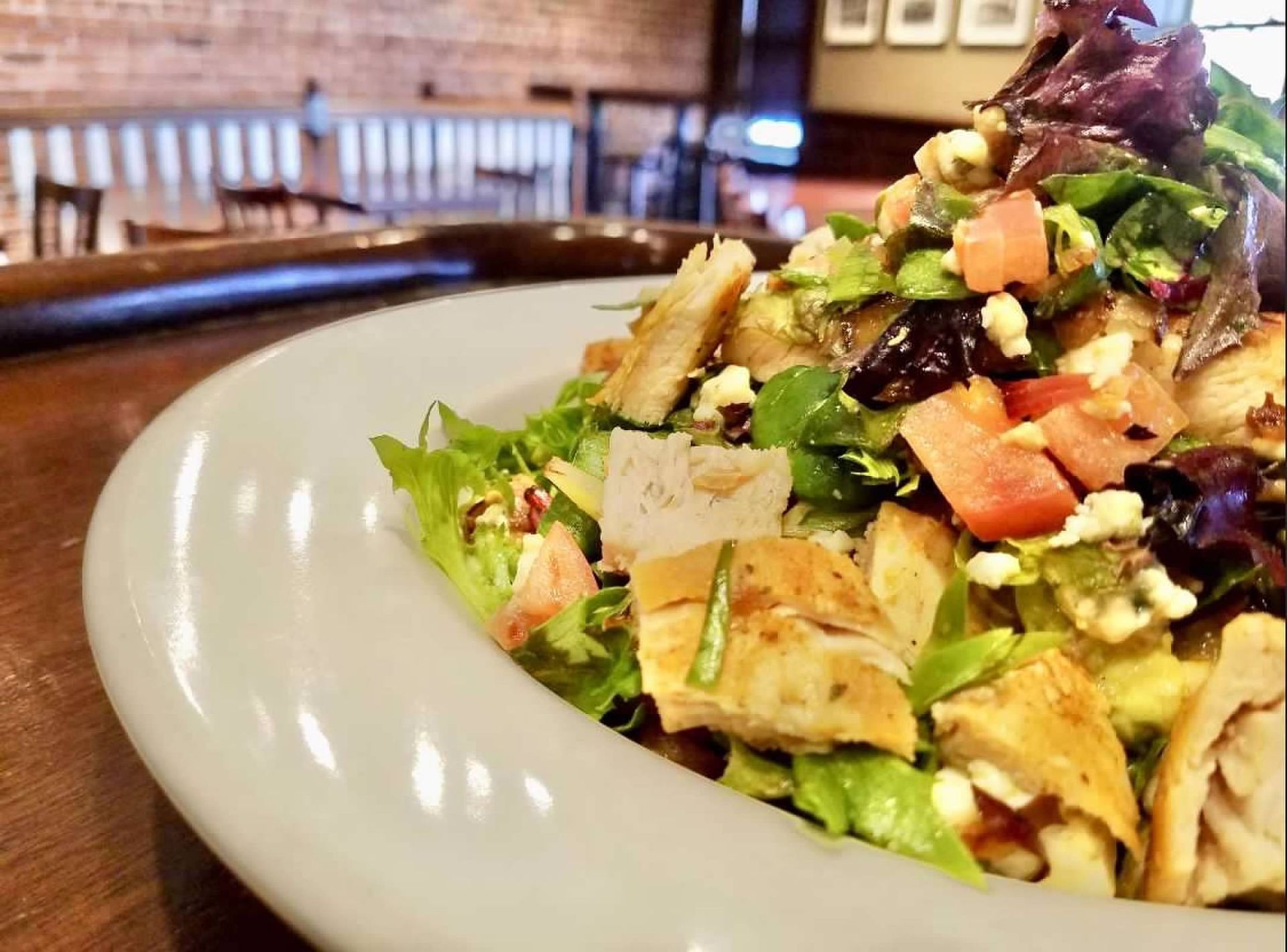 A close-up of a chicken salad with greens, tomatoes, and cheese served on a white plate in a restaurant setting.