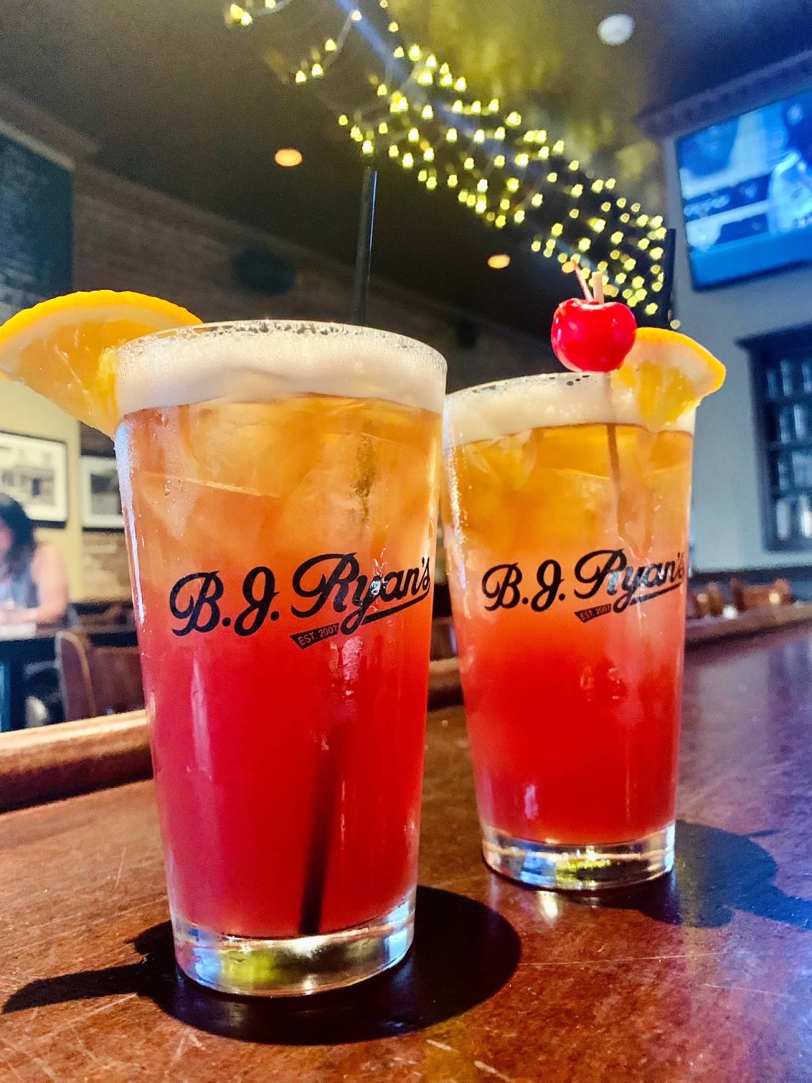 Two BJ Ryan's branded pint glasses containing layered, red-to-gold cocktails with ice, lemon slices, and a cherry garnish.