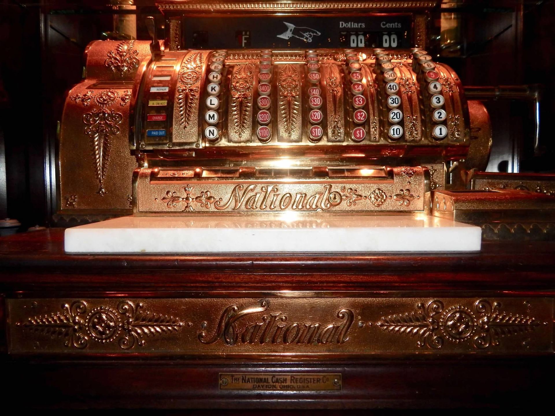 An antique, ornate bronze National brand cash register with a white marble surface sitting on a wooden base.