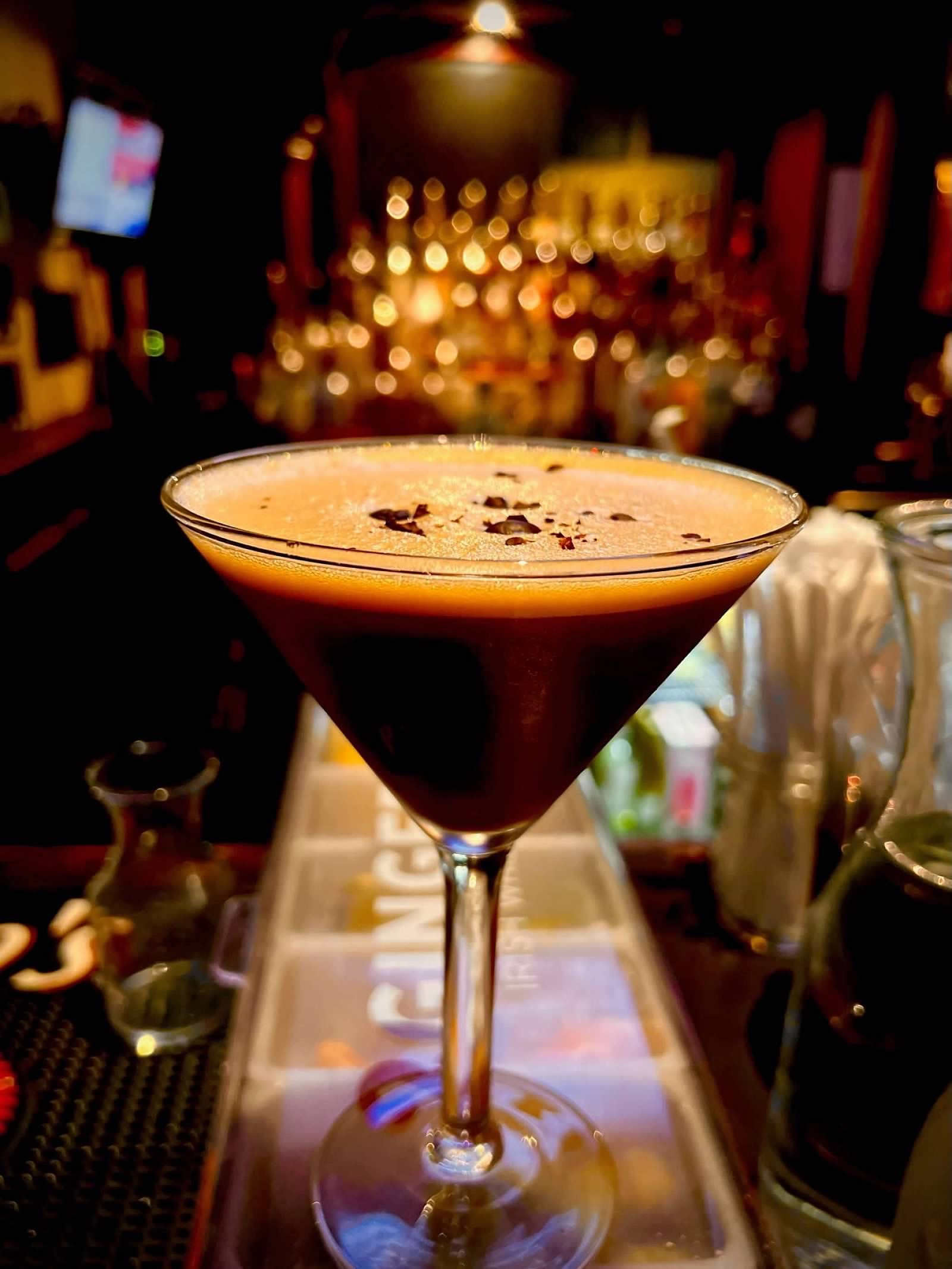 An espresso martini in a stemmed glass sits on a bar top with a blurred, warm-lit background of liquor bottles.