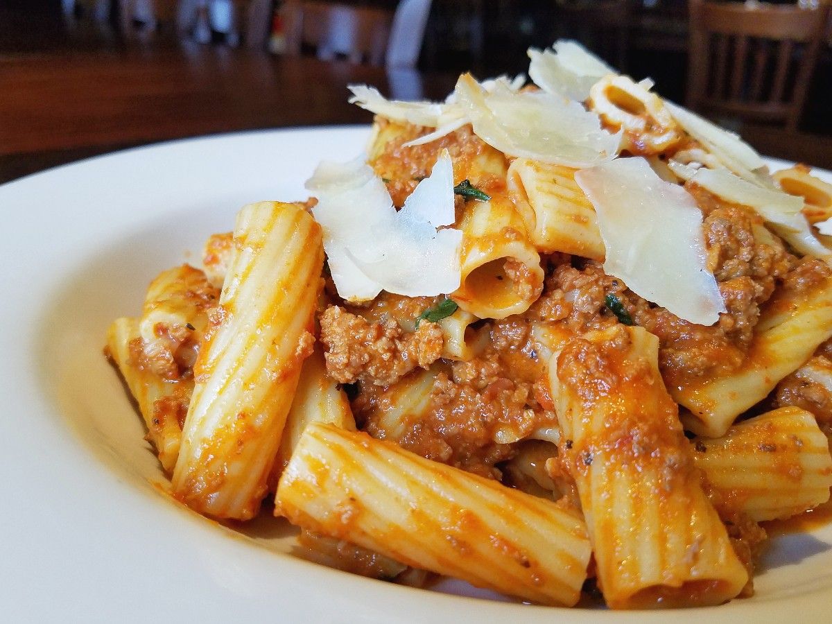 Rigatoni pasta with meat sauce, topped with thin shavings of parmesan cheese, served on a white plate.