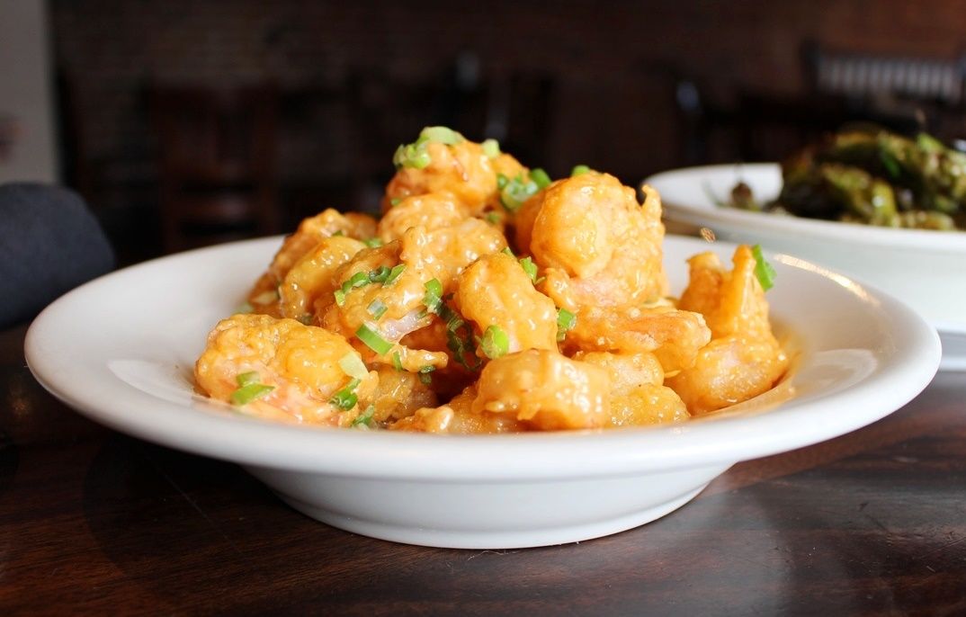A white bowl piled high with golden-fried popcorn shrimp topped with green onions on a dark wood table.