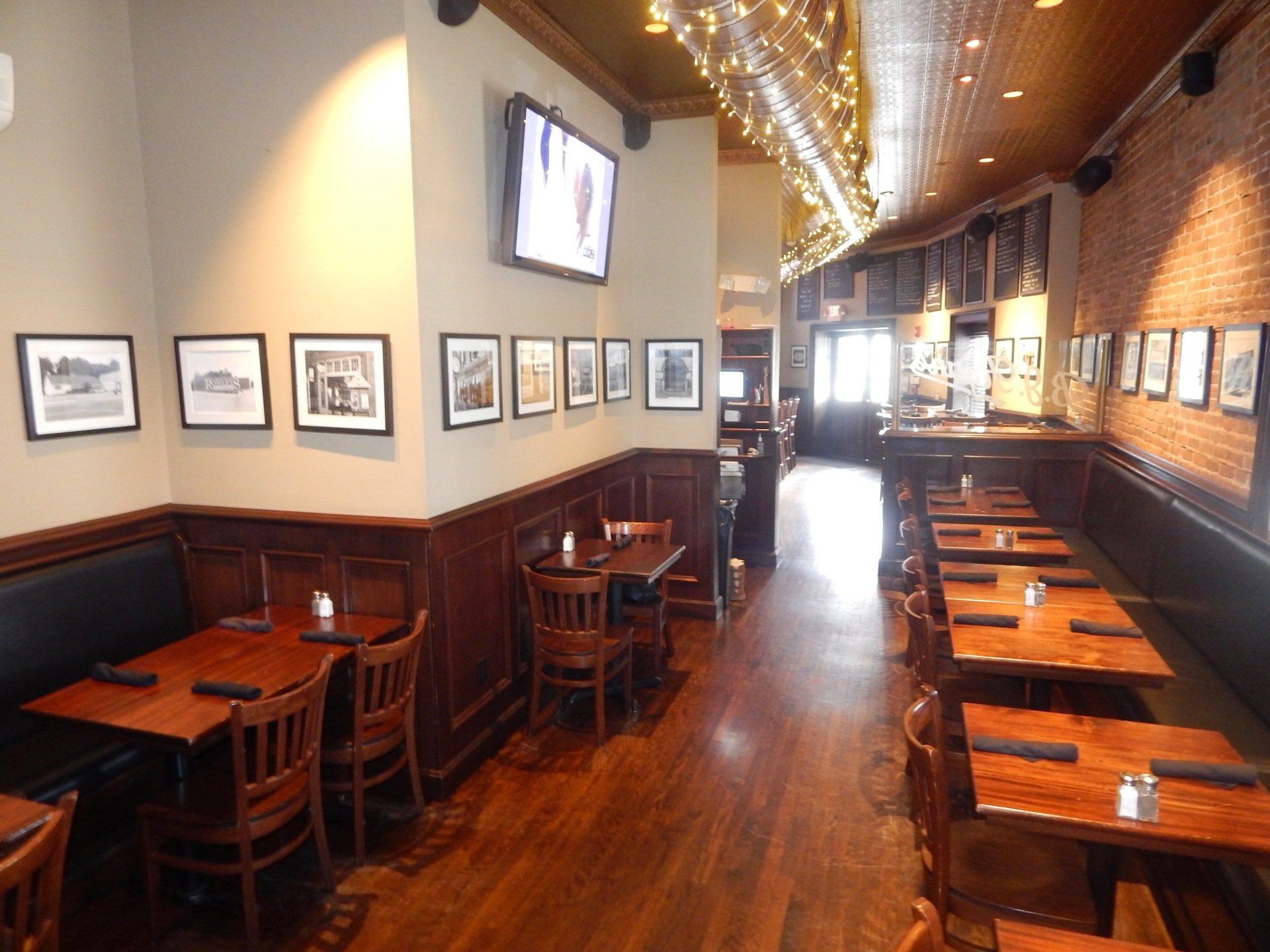 A restaurant interior with empty wooden tables, chairs, and booths, featuring brick walls and framed black-and-white photos.