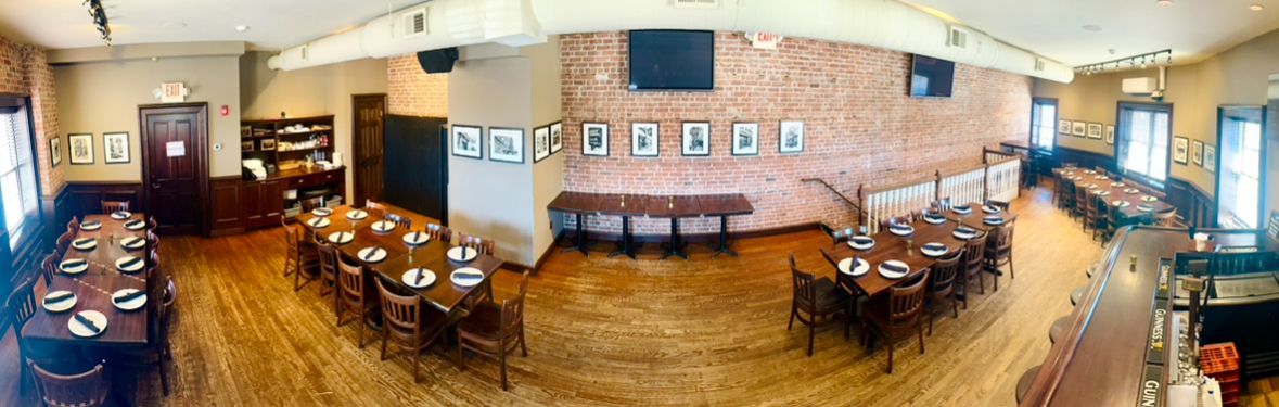 A panoramic view of a restaurant interior featuring wooden tables, chairs, and exposed brick walls lined with framed art.