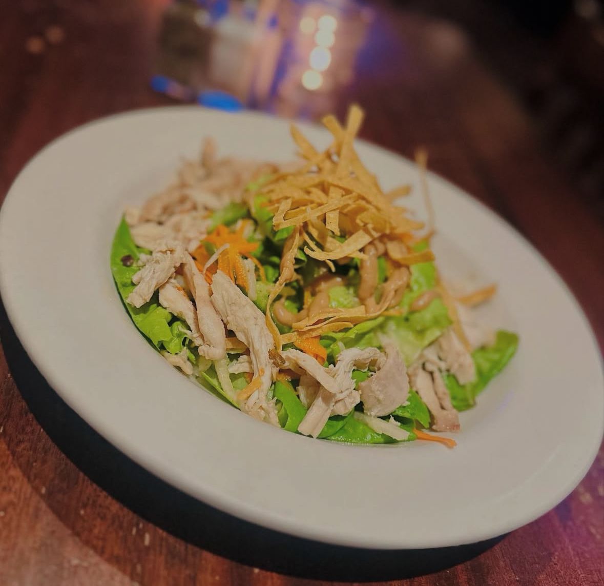 A chicken salad in a white bowl, topped with shredded carrots, green lettuce, and thin, crispy tortilla strips.