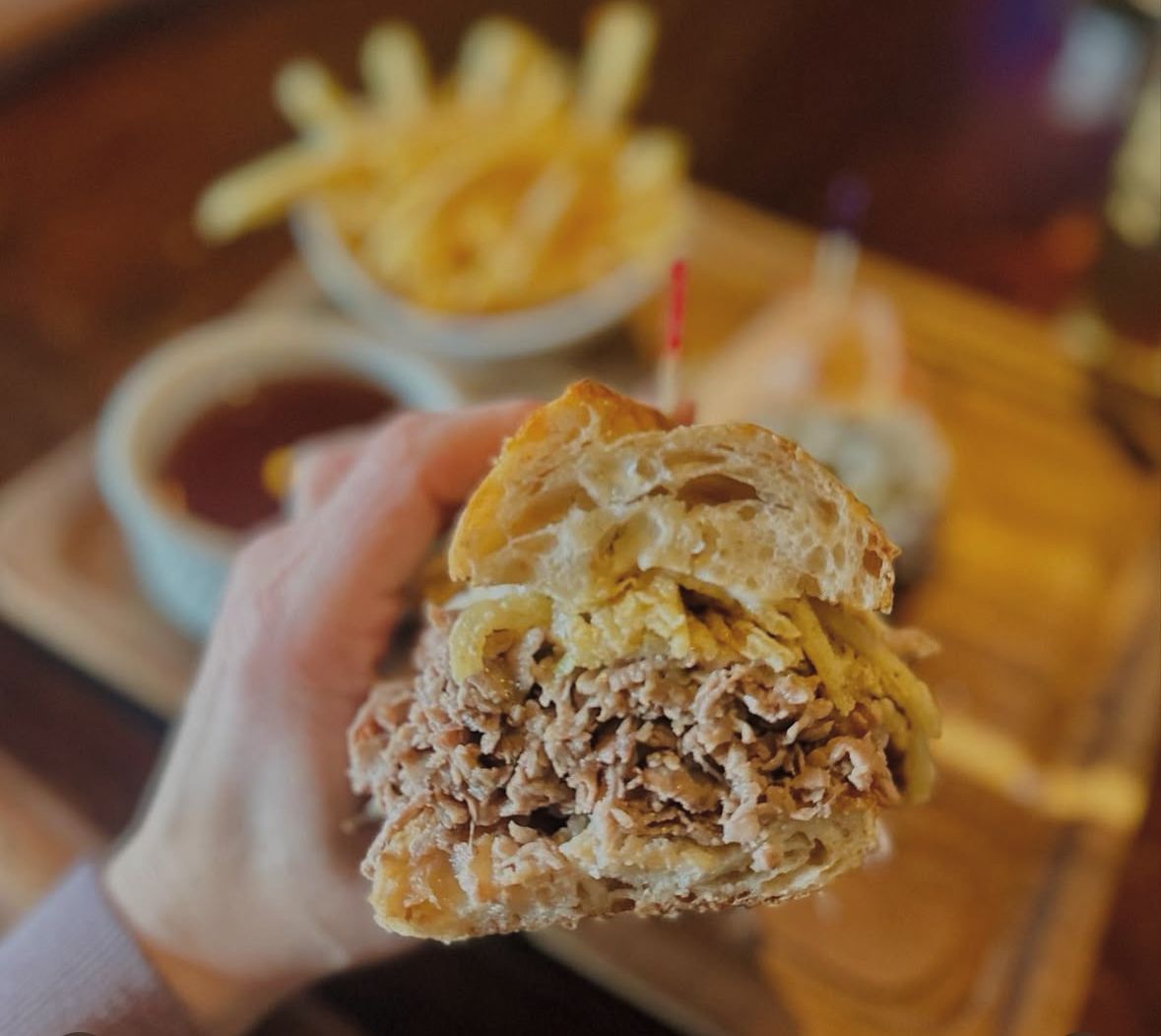 A hand holds a roast beef sandwich on a crusty roll, with a bowl of fries and dipping sauce on a wooden tray in the back.