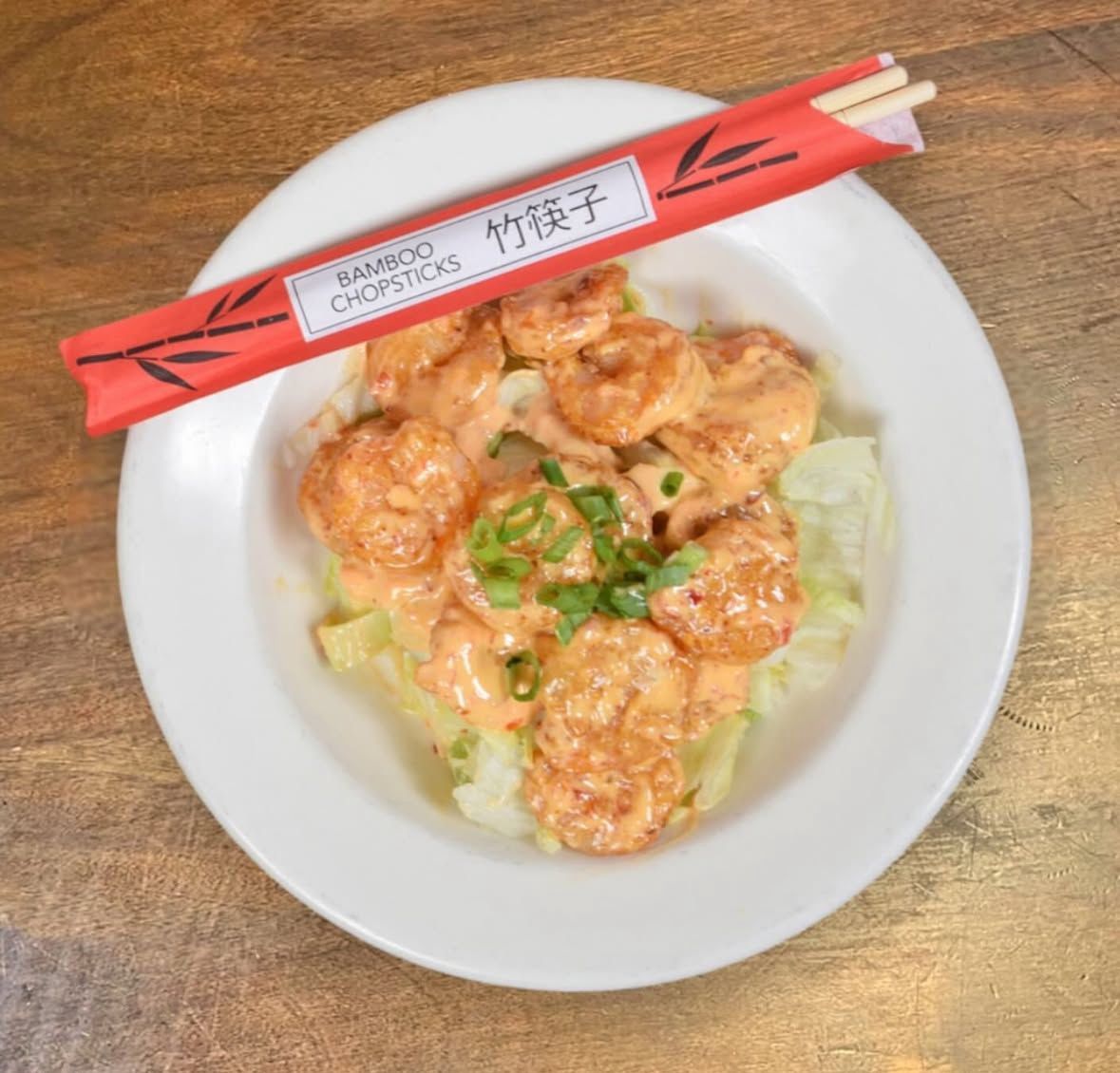 A bowl of shrimp tossed in a creamy sauce and topped with scallions, served with a red package of bamboo chopsticks.