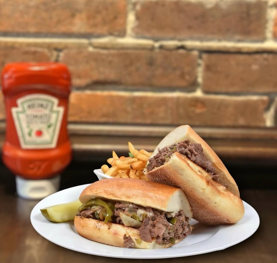 A Philly cheesesteak sandwich cut in half, a side of fries, and a pickle on a plate next to a bottle of Heinz ketchup.