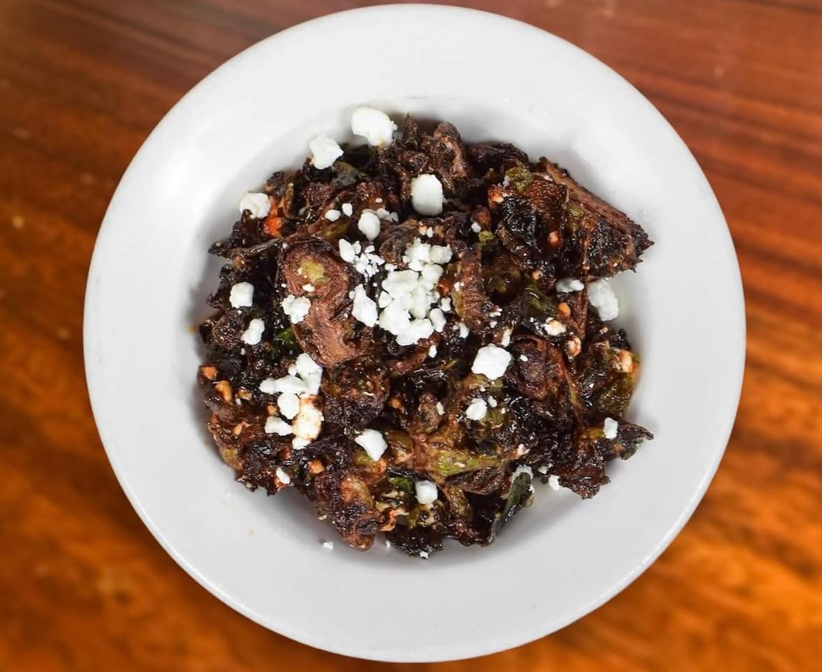 A white bowl filled with charred Brussels sprouts topped with crumbles of white cheese, set on a wooden table.