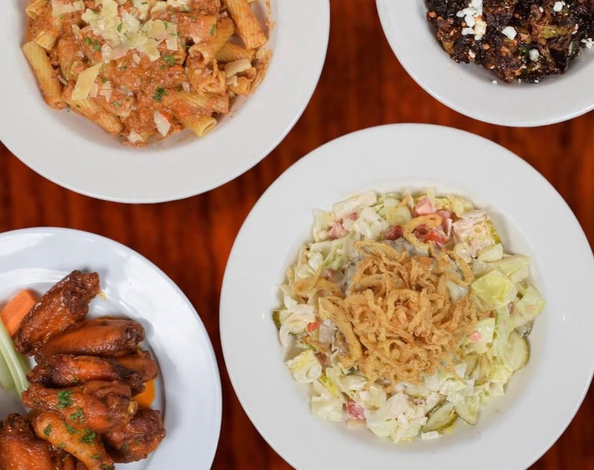 Top-down view of four white bowls on a wooden table, featuring pasta, crispy fried food, chicken wings, and a salad.