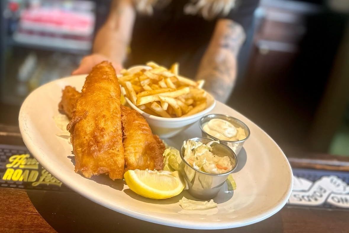 A plate of battered fish and chips served with lemon wedges and two small ramekins of sauce, held by someone.