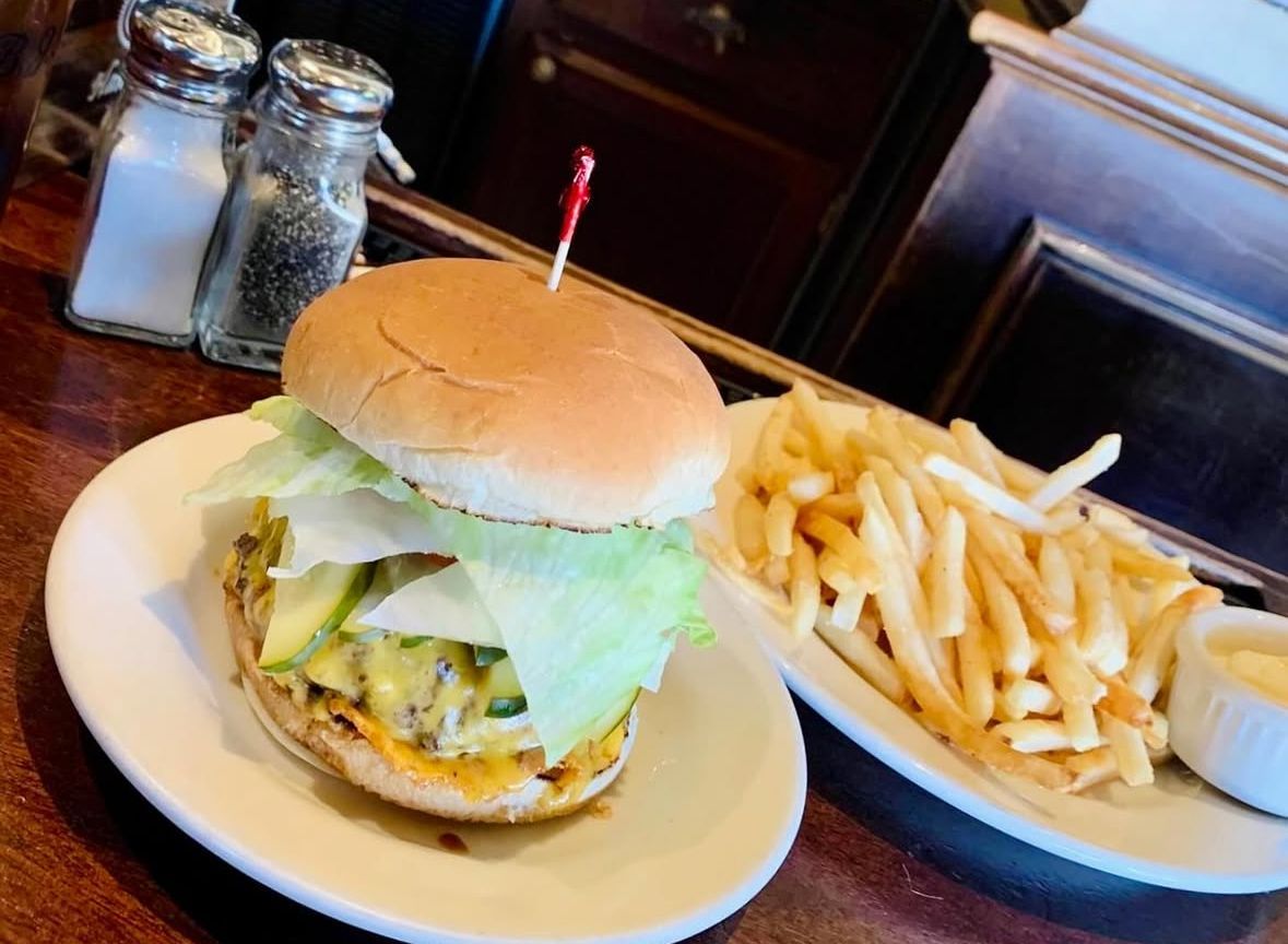 A cheeseburger topped with lettuce and a toothpick, served with a side of french fries and a small dish of sauce.