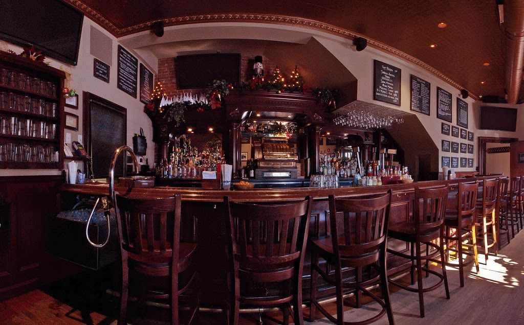 A dark, wooden bar with a row of empty chairs, glass shelves, and a polished counter in a cozy, dimly lit interior.