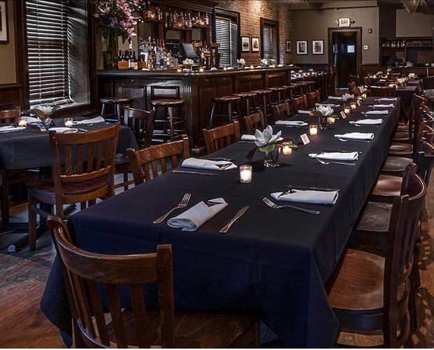 A long table set for dining with dark blue tablecloths, candles, and wooden chairs in a restaurant with a bar background.
