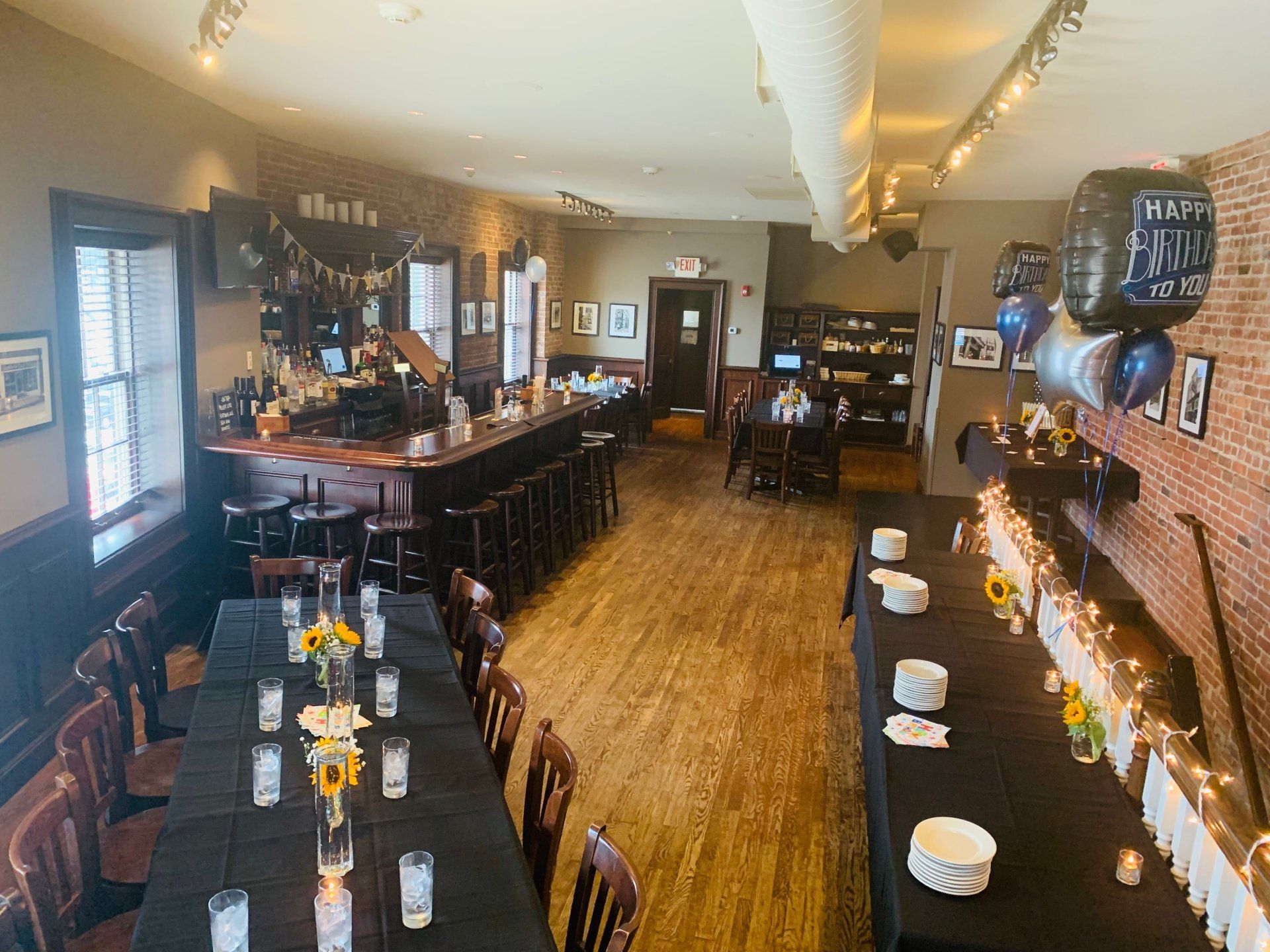 An indoor dining area featuring long tables with black tablecloths set for a party, a bar, and birthday balloons.