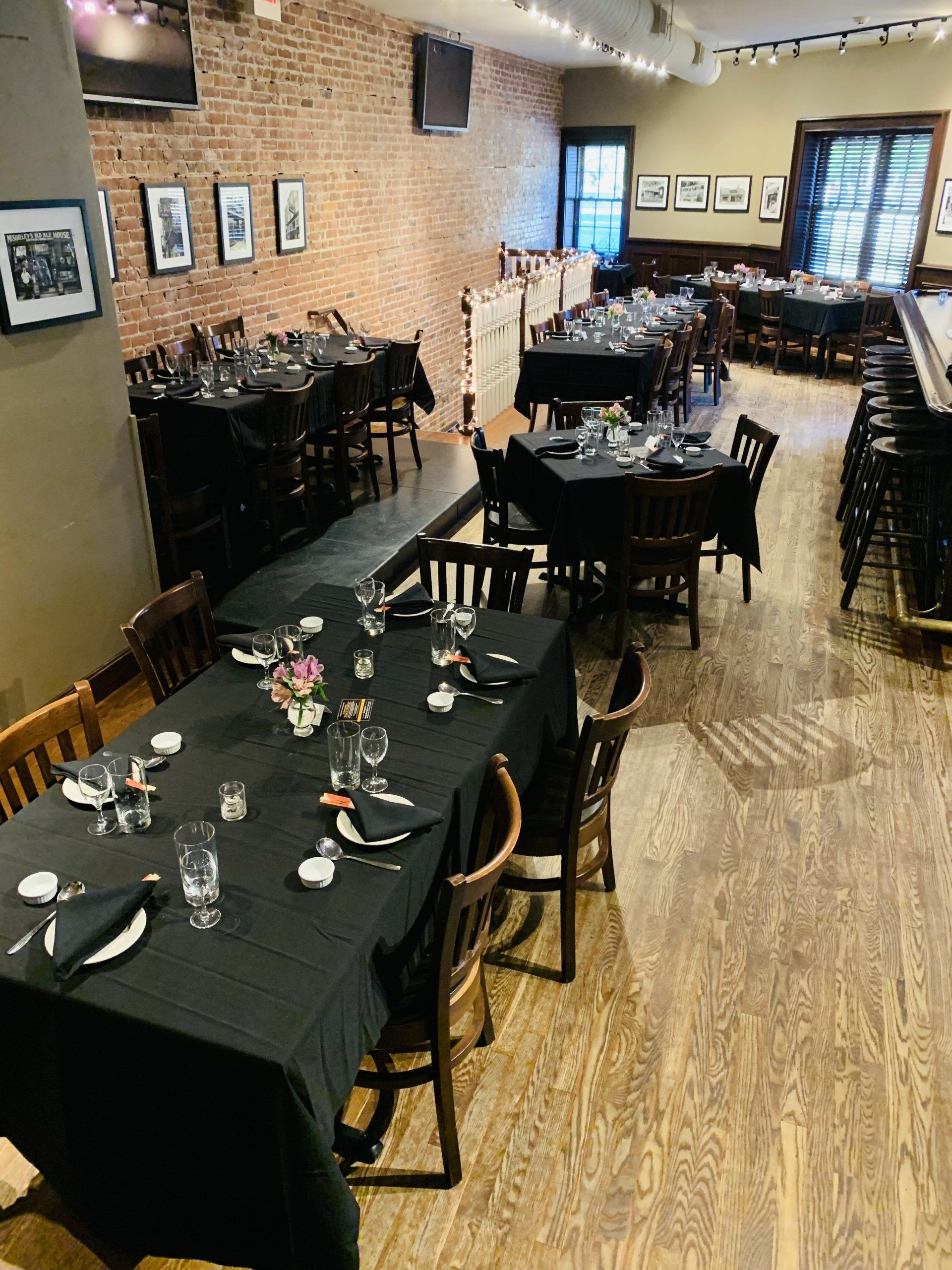 A dining area with multiple tables covered in black tablecloths arranged on a hardwood floor by a rustic brick wall.
