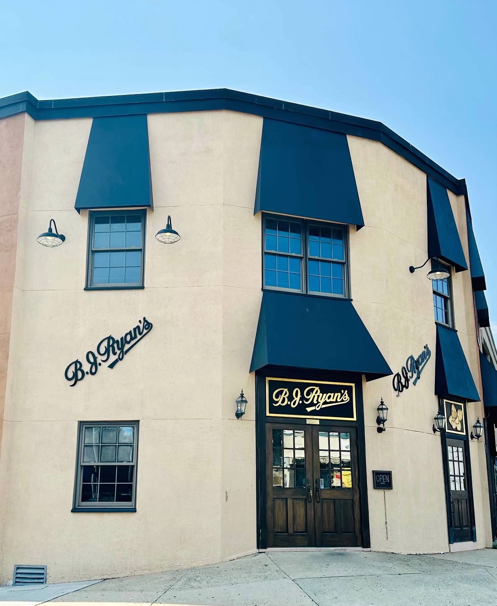 A corner view of the B.J. Ryan’s establishment, featuring a tan building with dark blue window awnings and wooden doors.