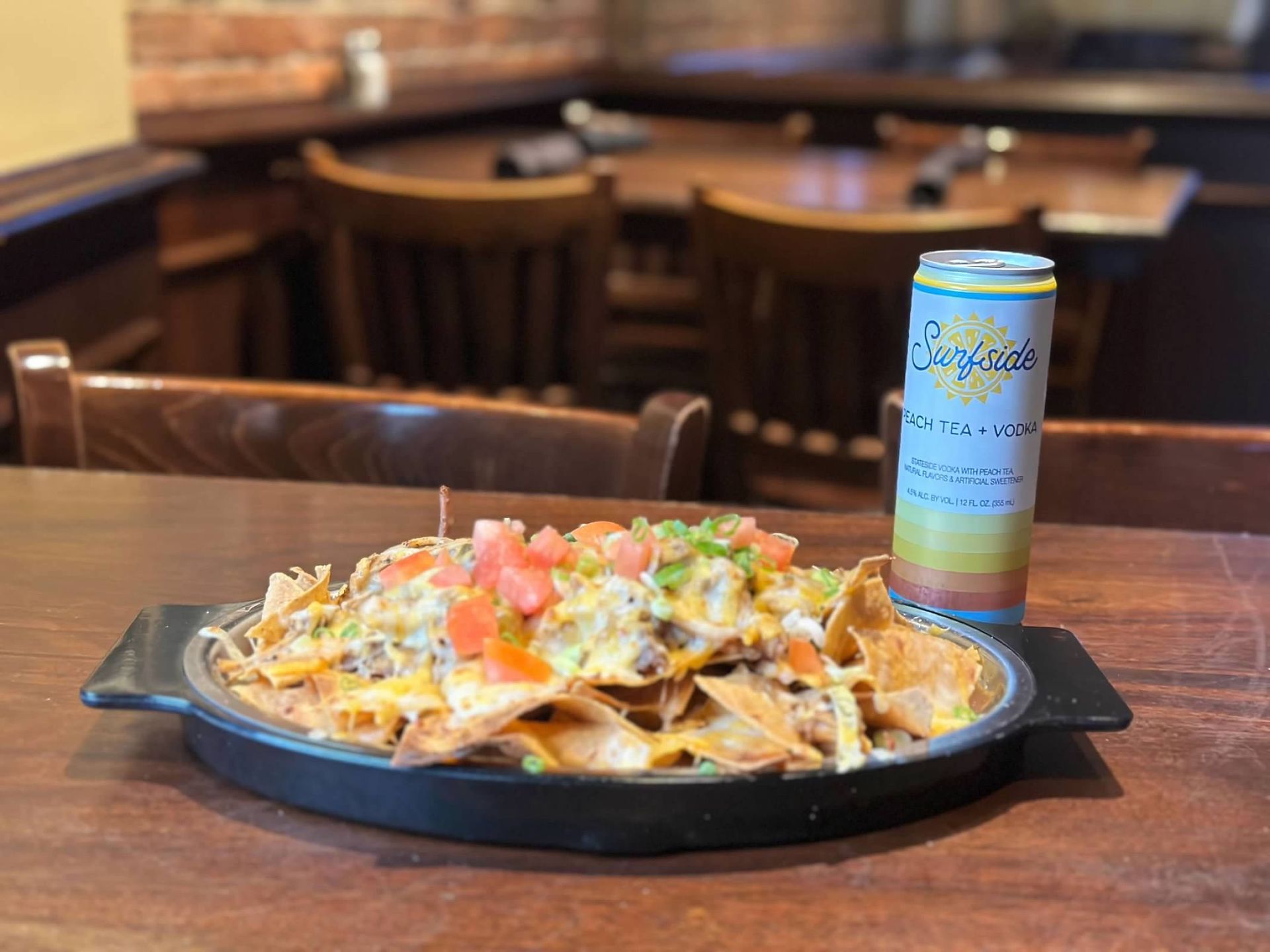 A plate of loaded nachos topped with cheese and tomatoes sits on a wooden table next to a canned beverage.