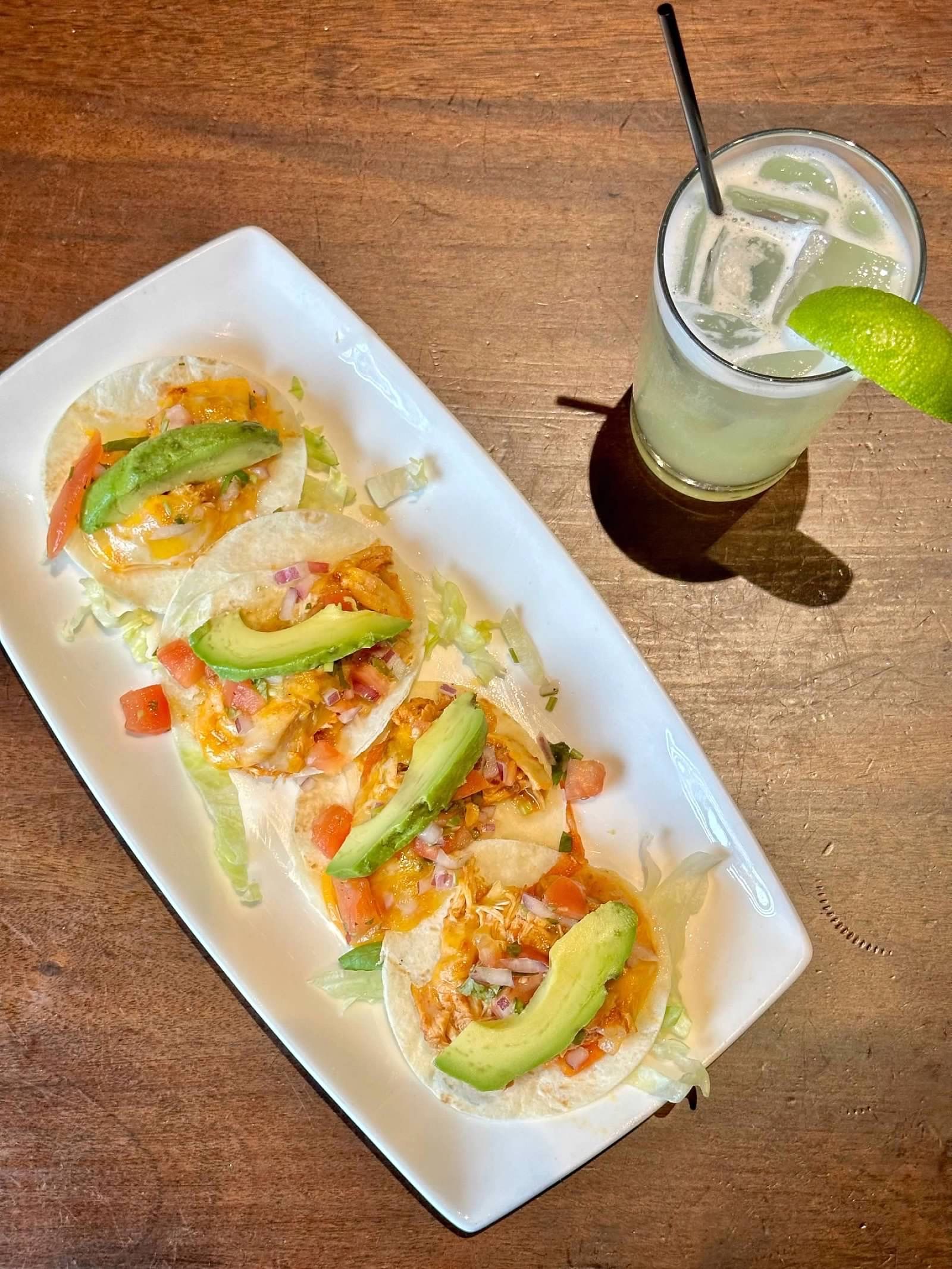 Four tacos topped with avocado, tomato, and lettuce on a rectangular plate next to a margarita with a lime wedge.