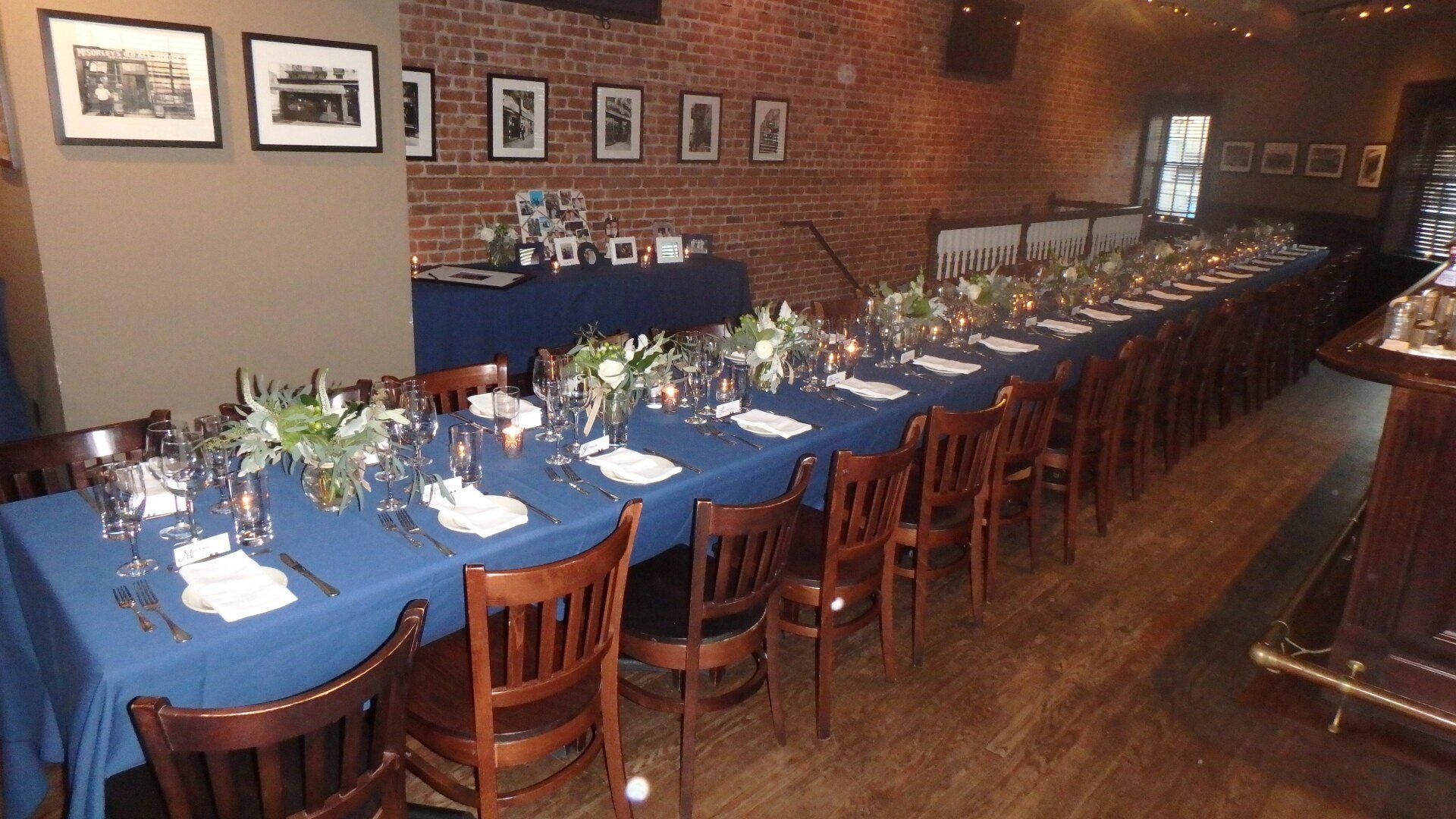 A long dining table set with blue tablecloths, white napkins, and floral centerpieces in a rustic room with brick walls.