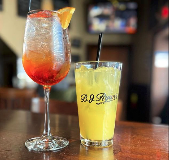 Two cocktails on a wooden bar top: a vibrant orange Aperol spritz in a wine glass and a yellow drink in a B.J. Ryan's glass.
