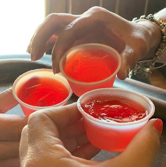 Three hands holding small plastic cups filled with bright red gelatin shots in a toast.