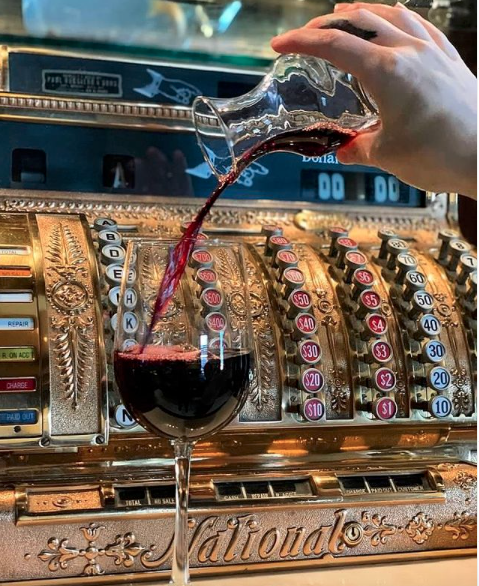 Red wine being poured from a small glass carafe into a stemmed wine glass in front of an antique brass cash register.