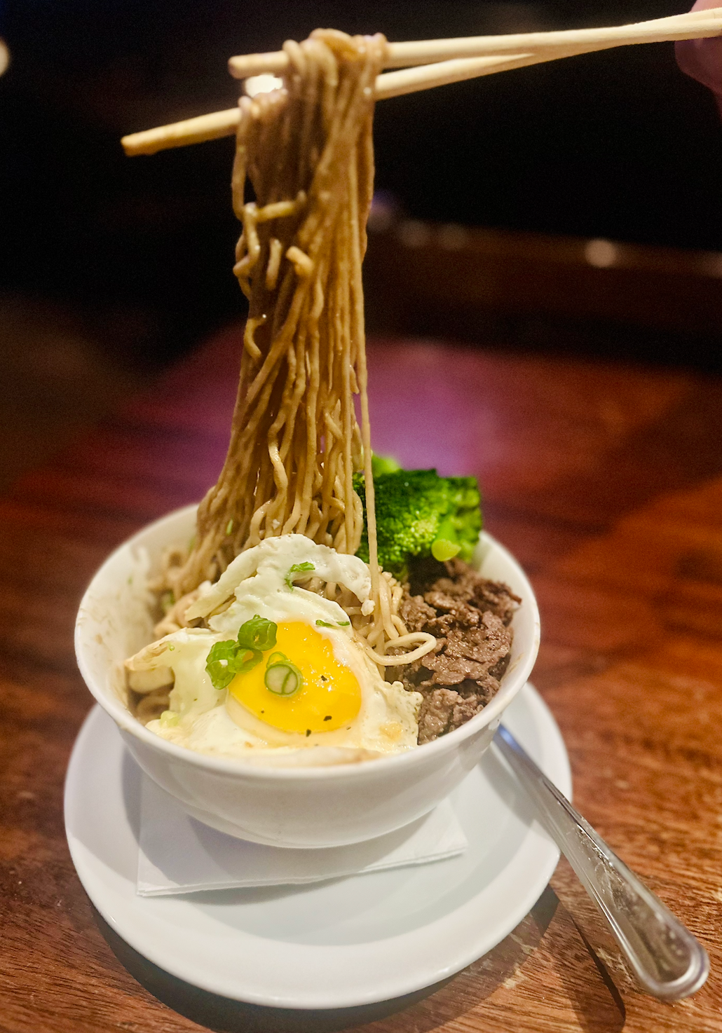 Chopsticks hold a bundle of noodles over a bowl containing a fried egg, broccoli, and ground meat on a wooden table.