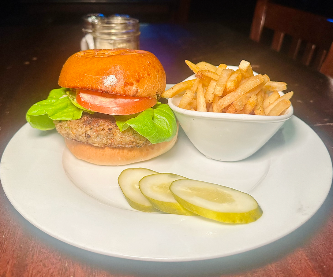 A burger with lettuce and tomato, a side of fries, and three pickle slices on a white plate on a dark wooden table.
