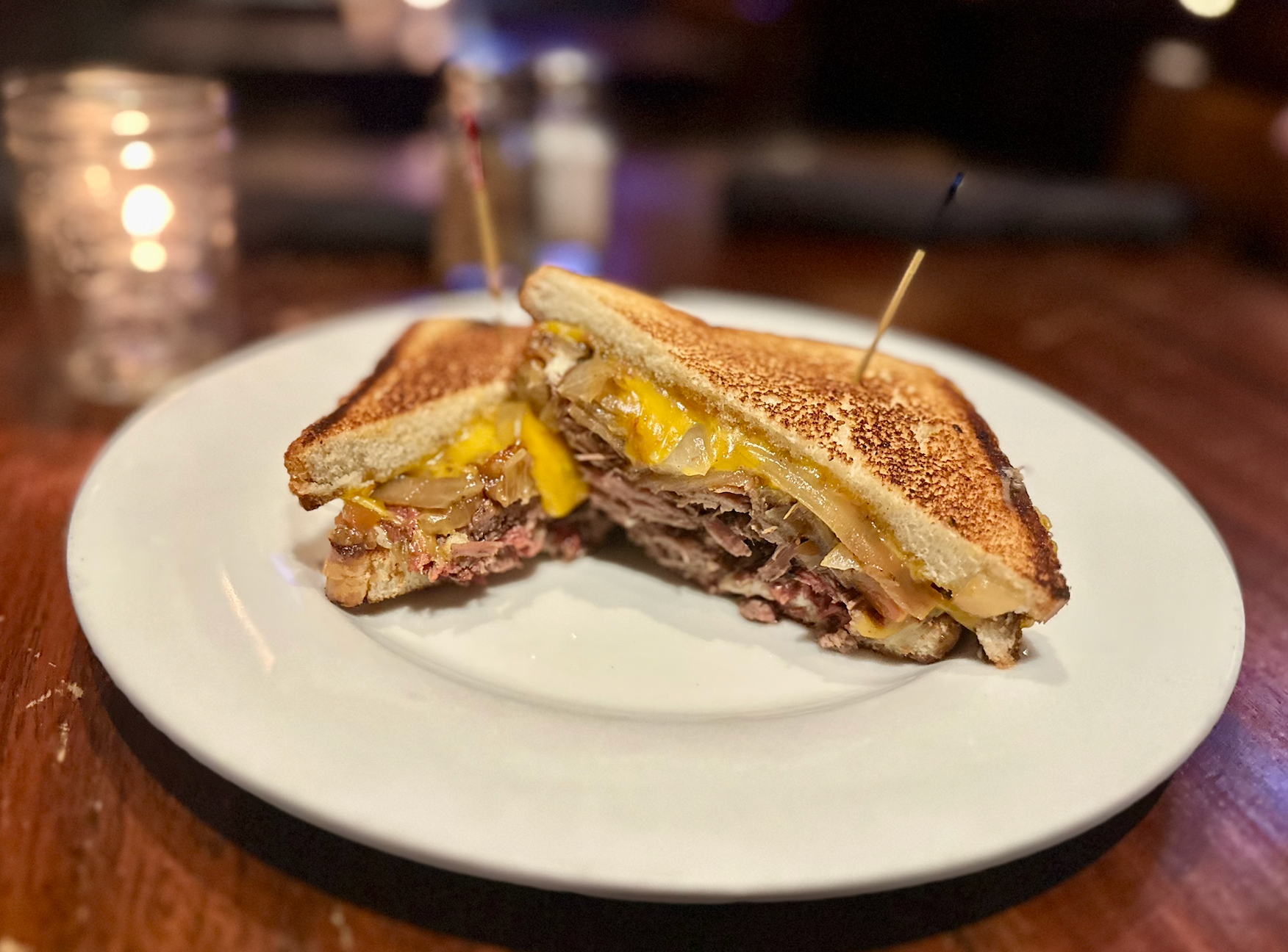 A grilled sandwich cut in half on a white plate, served with toothpicks on a dark wooden table with a blurred background.