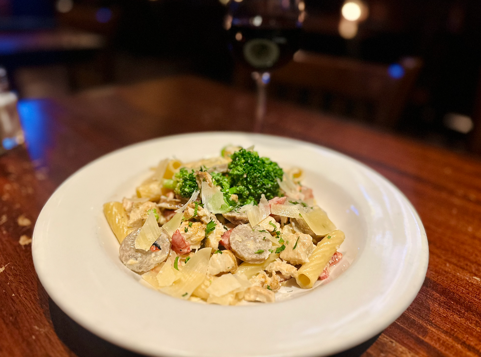 A bowl of penne pasta with sausage and herbs in a creamy sauce, served with a glass of red wine on a wooden table.