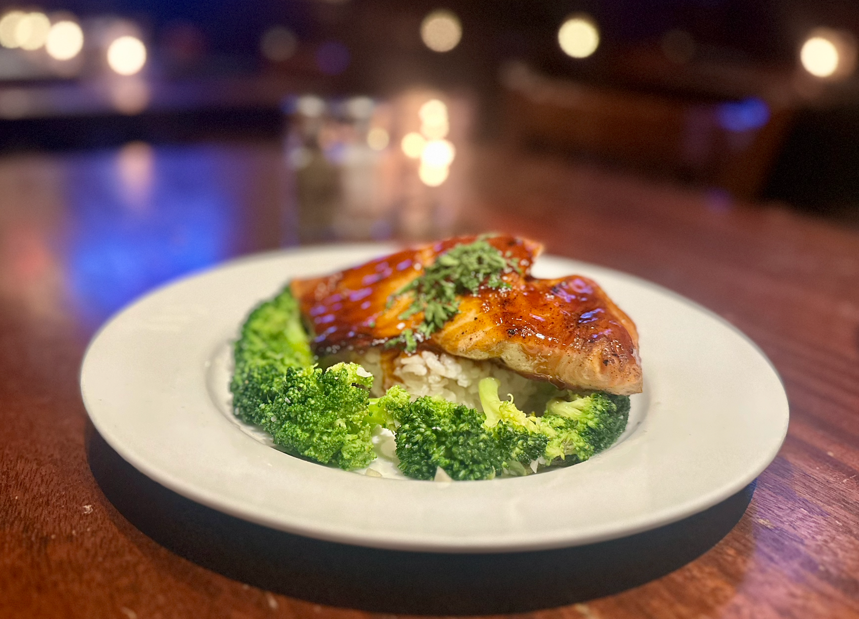 A glazed fish fillet topped with herbs, served over rice with steamed broccoli on a white plate on a dark table.