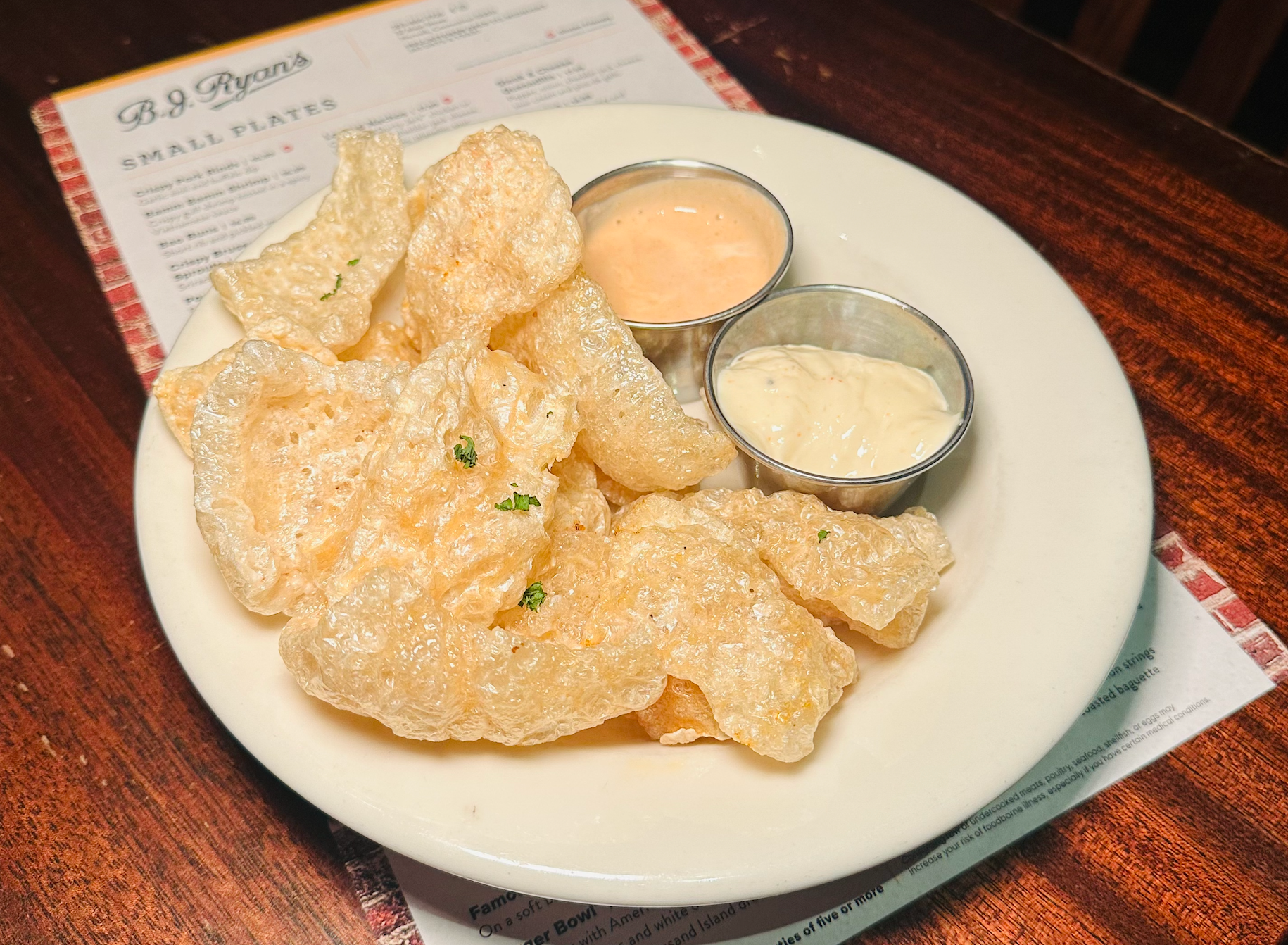 A plate of crispy pork rinds with two small metal cups of dipping sauces, placed on a wooden restaurant table.