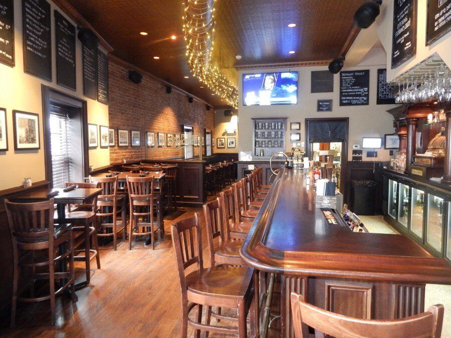 A cozy tavern interior featuring a long wooden bar, high stools, a brick wall, and menu boards hanging on the wall.