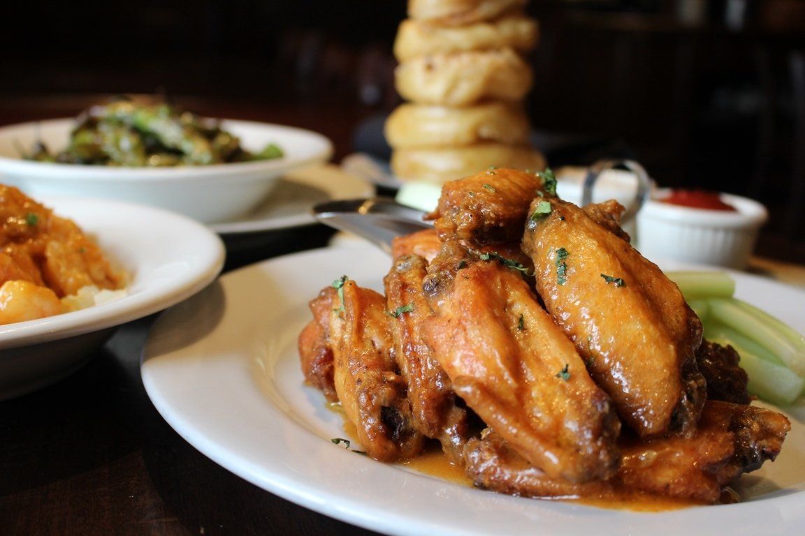 A plate of saucy chicken wings with celery sticks, flanked by plates of onion rings and green beans on a dark table.