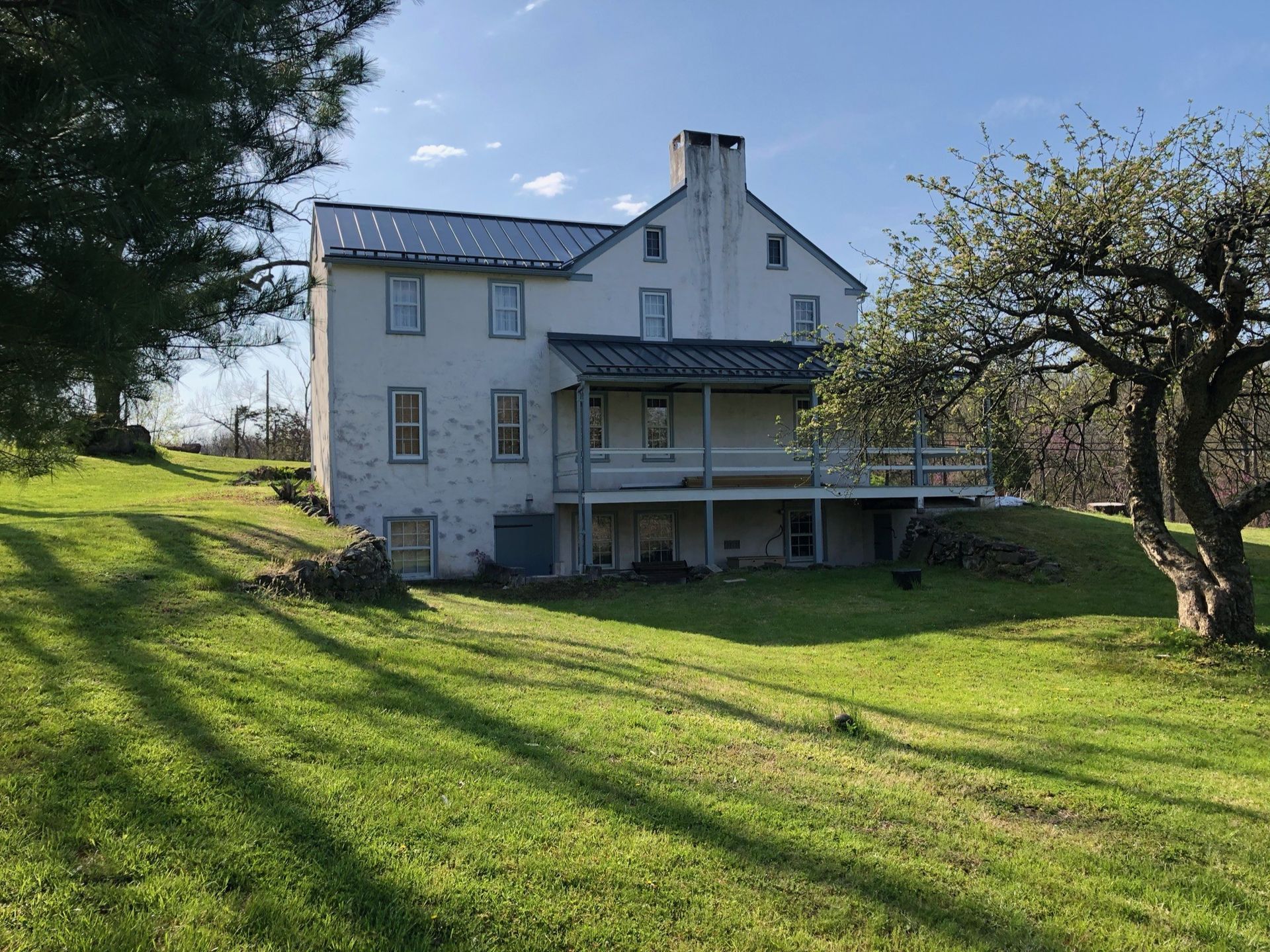 Two-story white farmhouse with porch, chimney, and metal roof, on a grassy hill, under a blue sky.