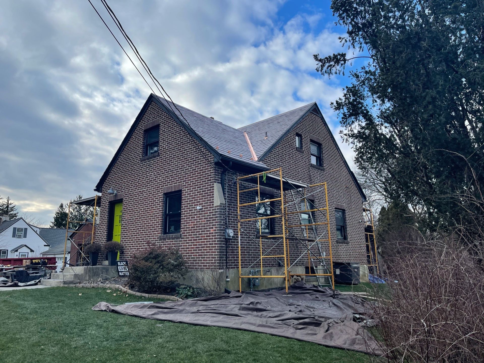 House with fire damage, scaffolding, and tarp on the lawn under a cloudy sky.