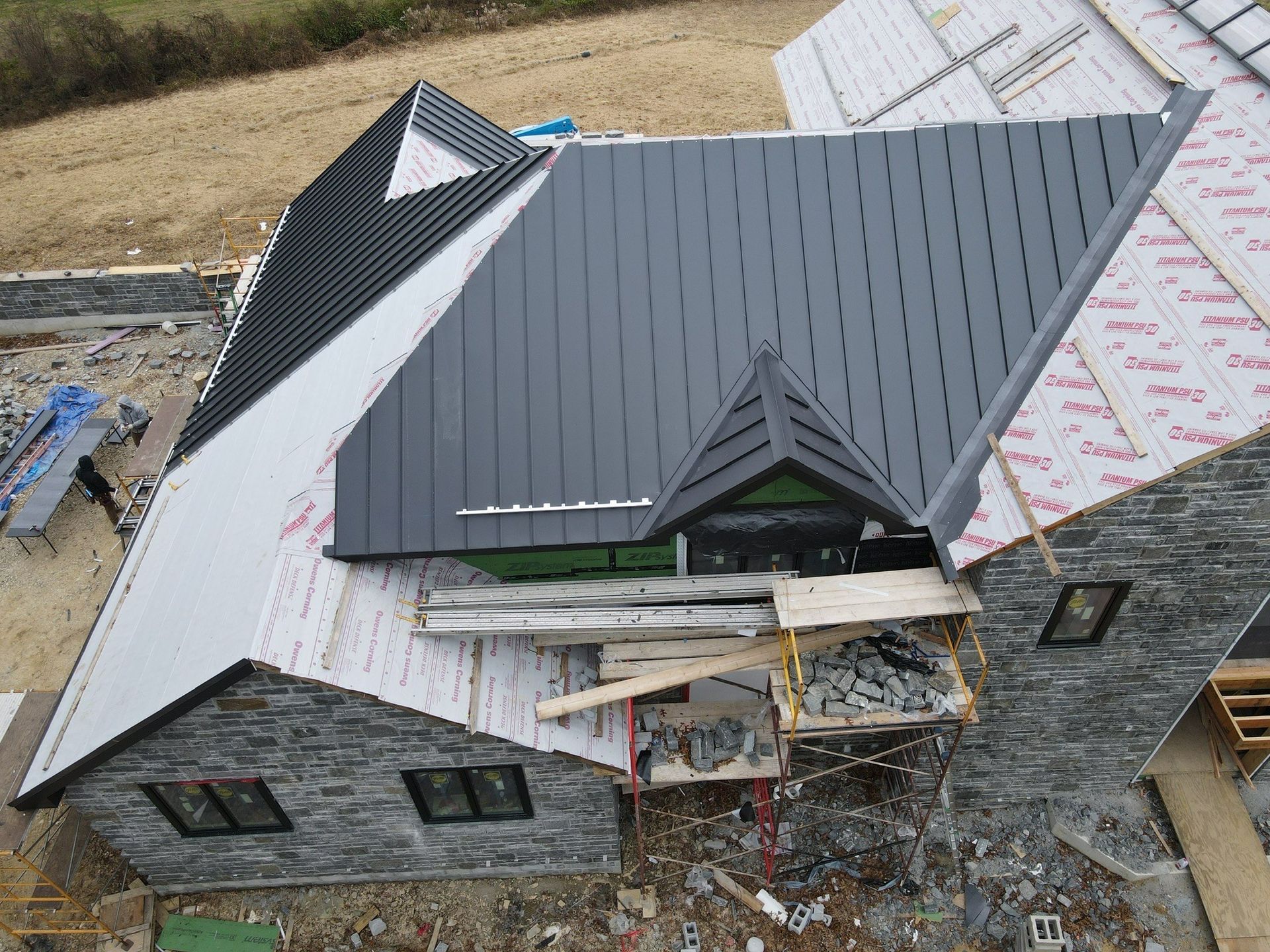 Overhead view of a house under construction with a dark gray metal roof, light gray roof sections, and stone siding.