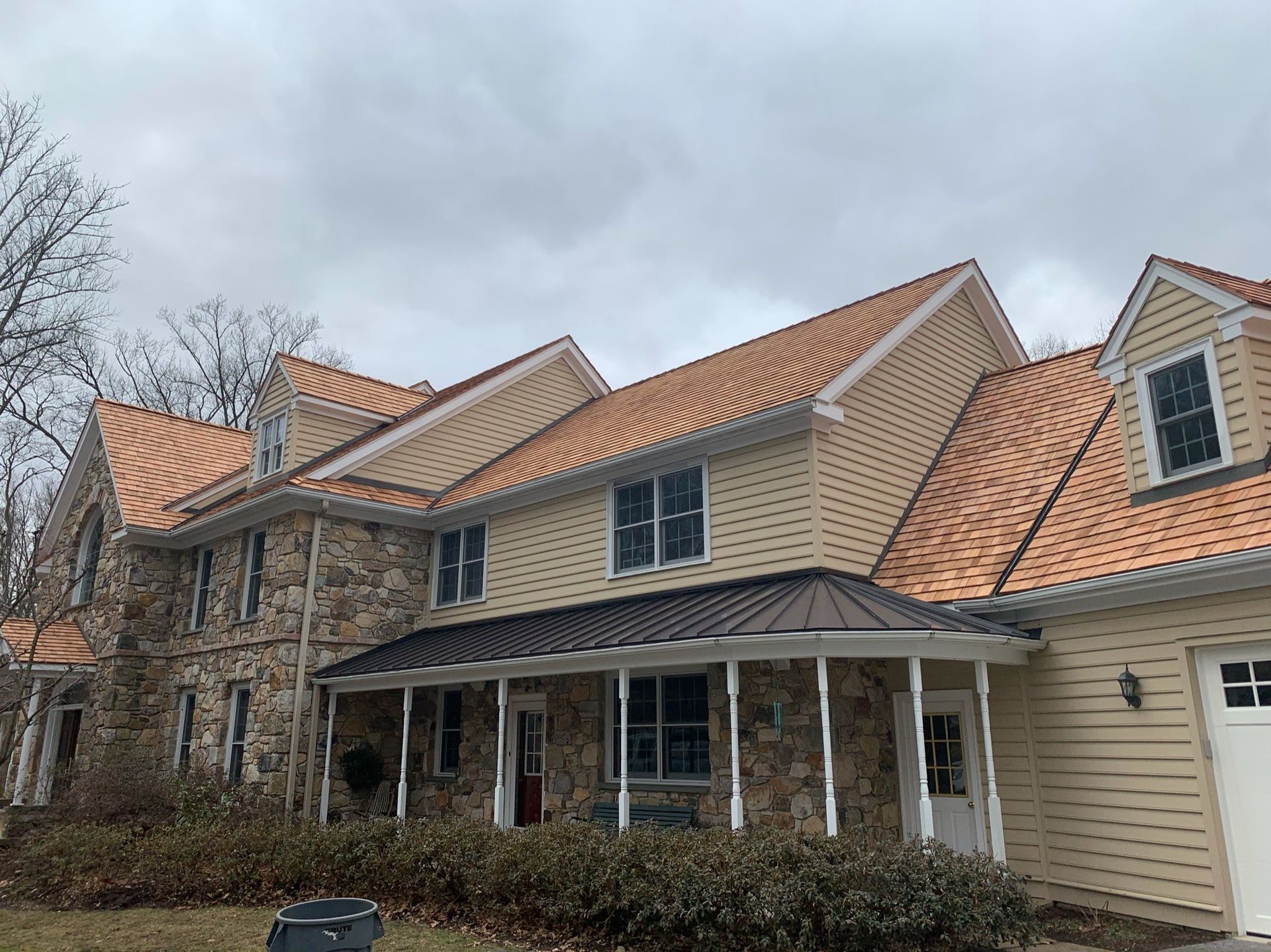Two-story house with stone and yellow siding, tan roof. Porch with dark roof and white columns. Overcast sky.