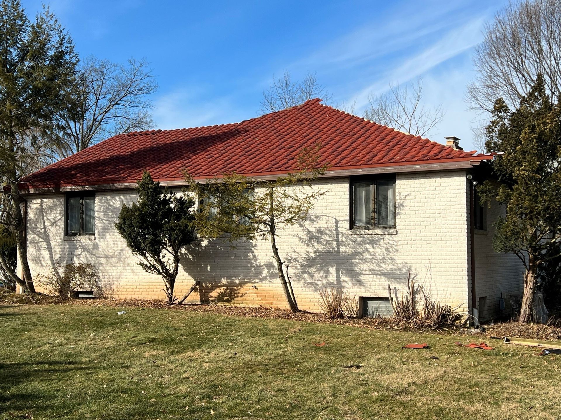 A one-story brick house with a red tiled roof, windows, and small trees in front.