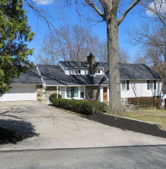 Suburban house with a driveway. White siding, black roof, and a large tree in the front yard.