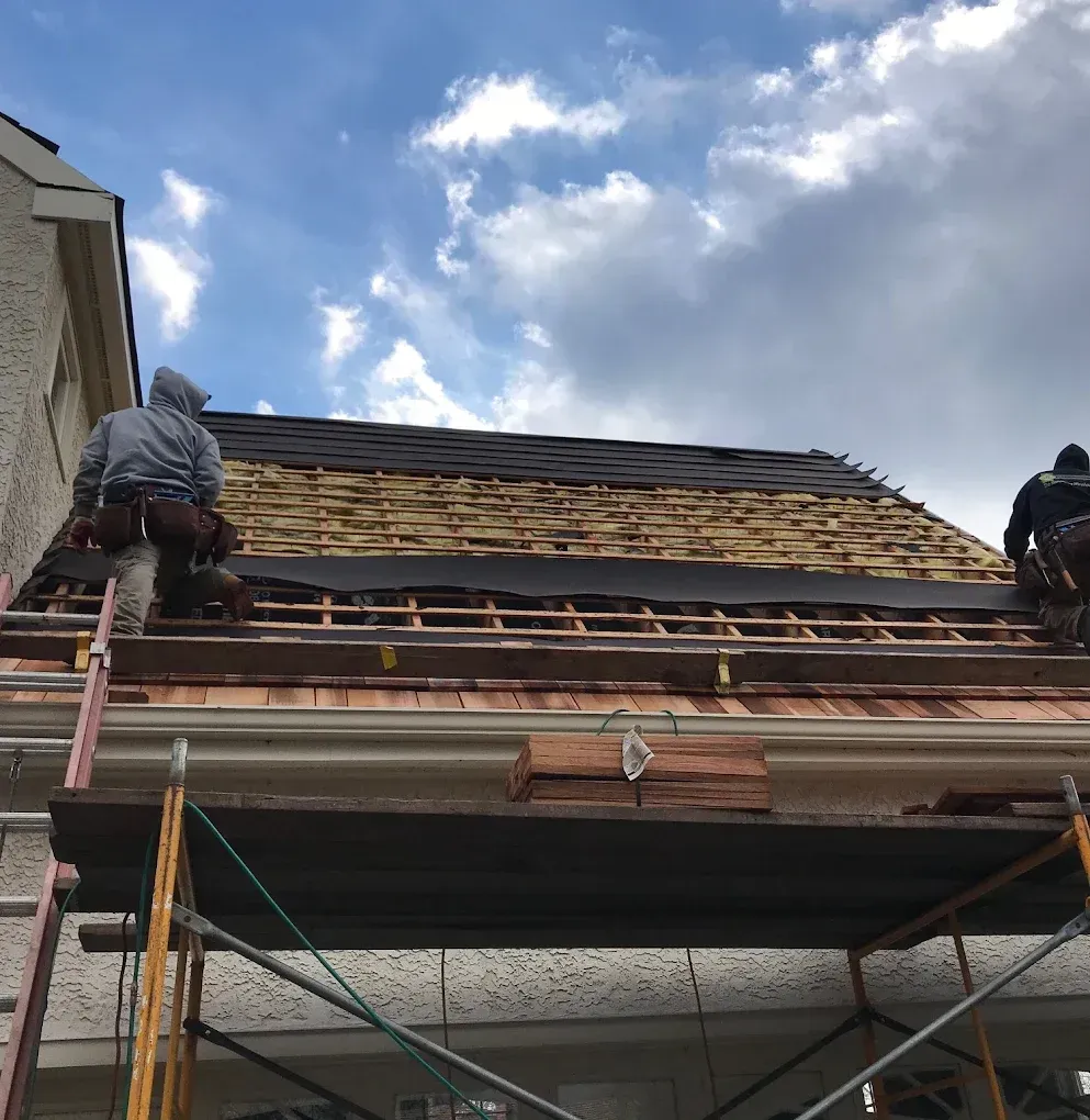 Two roofers replacing shingles on a house roof under a cloudy sky.