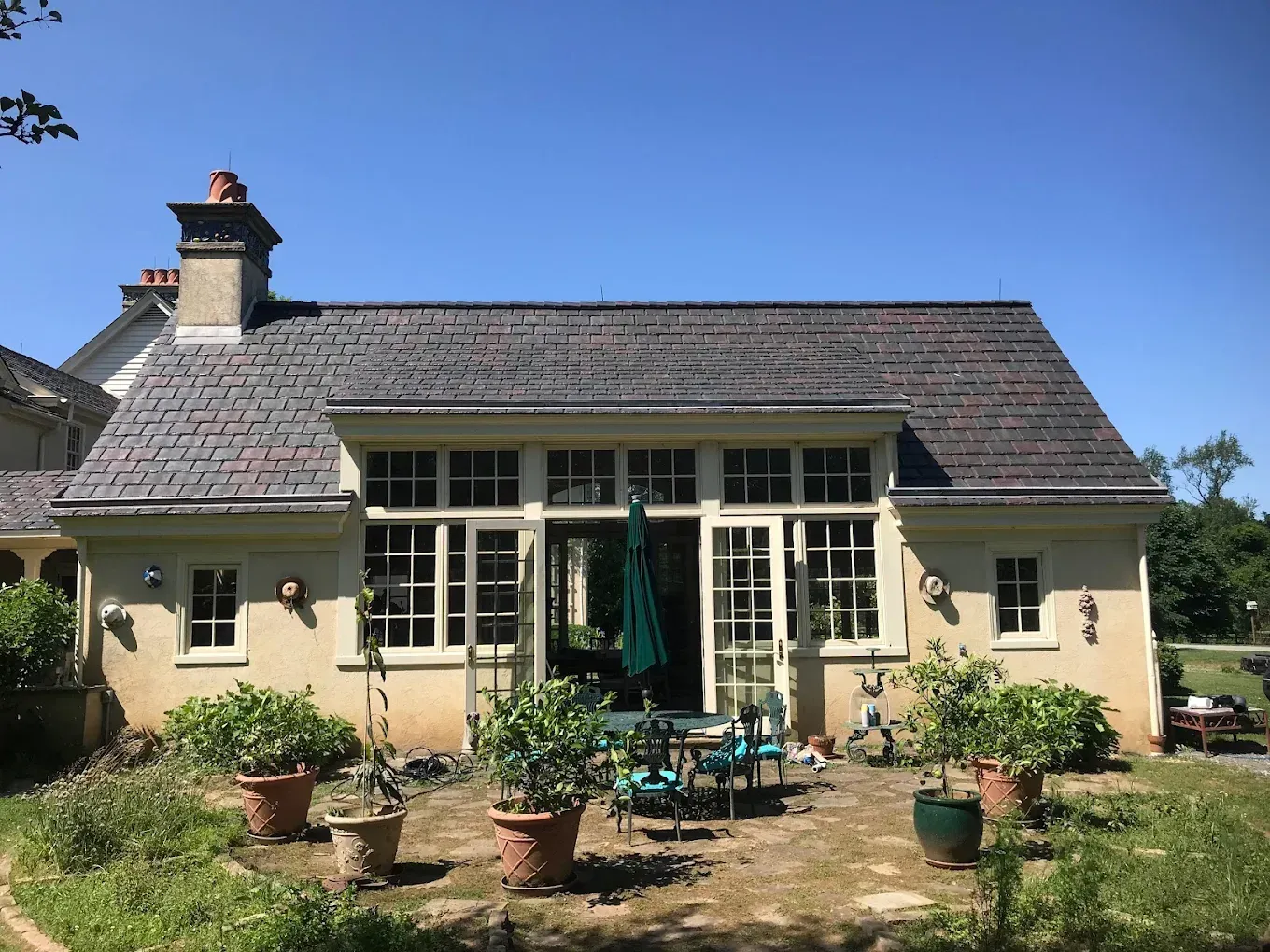 Back of a cream-colored house with a dark gray tiled roof, french doors, windows, and potted plants on a patio.