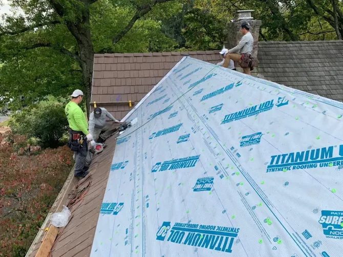 Two roofers installing roofing material on a house, working at the top of a roof.