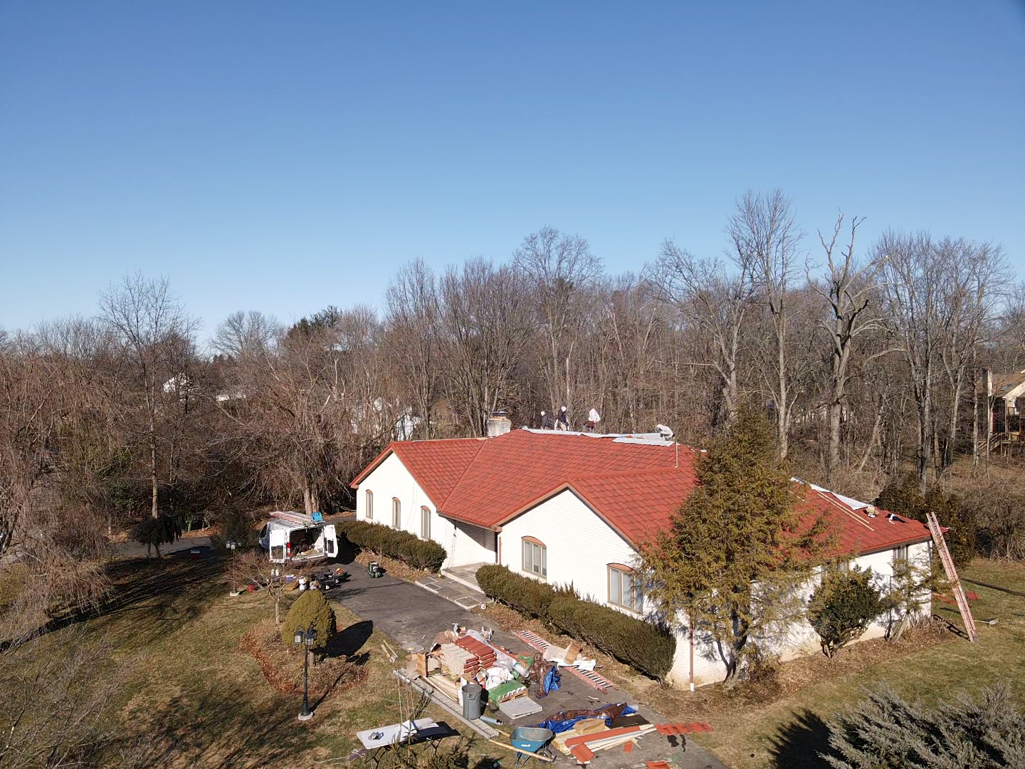 House with red tile roof, white walls, and a driveway in a wooded area on a sunny day.