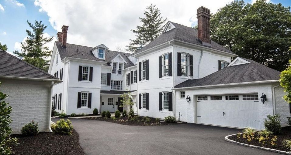 White two-story house with black shutters, attached garage, and black asphalt driveway against a cloudy sky.
